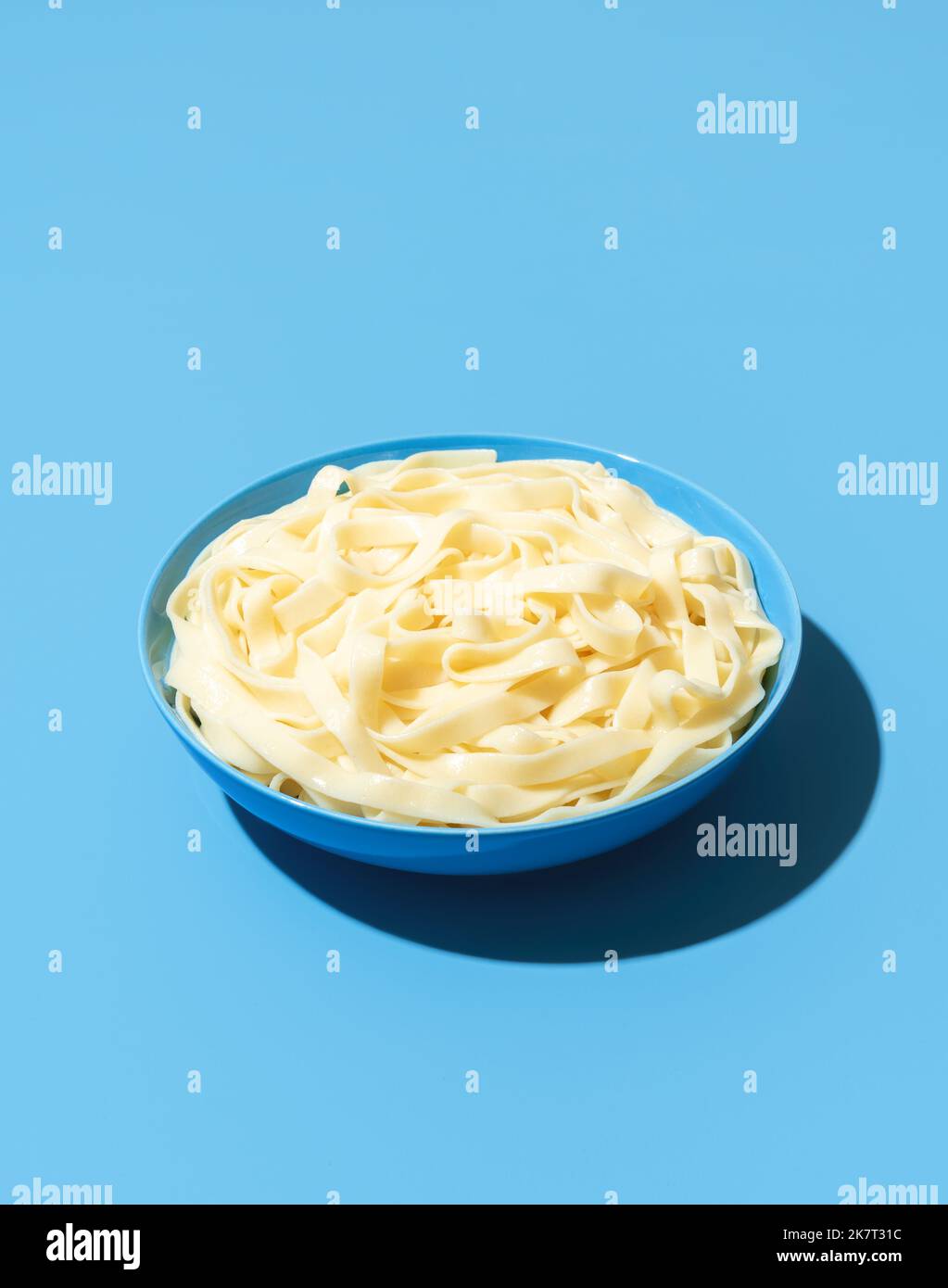 Close-up with a bowl full of boiled tagliatelle pasta isolated on a blue background. Bowl with plain noodles minimalist on a colorful table Stock Photo