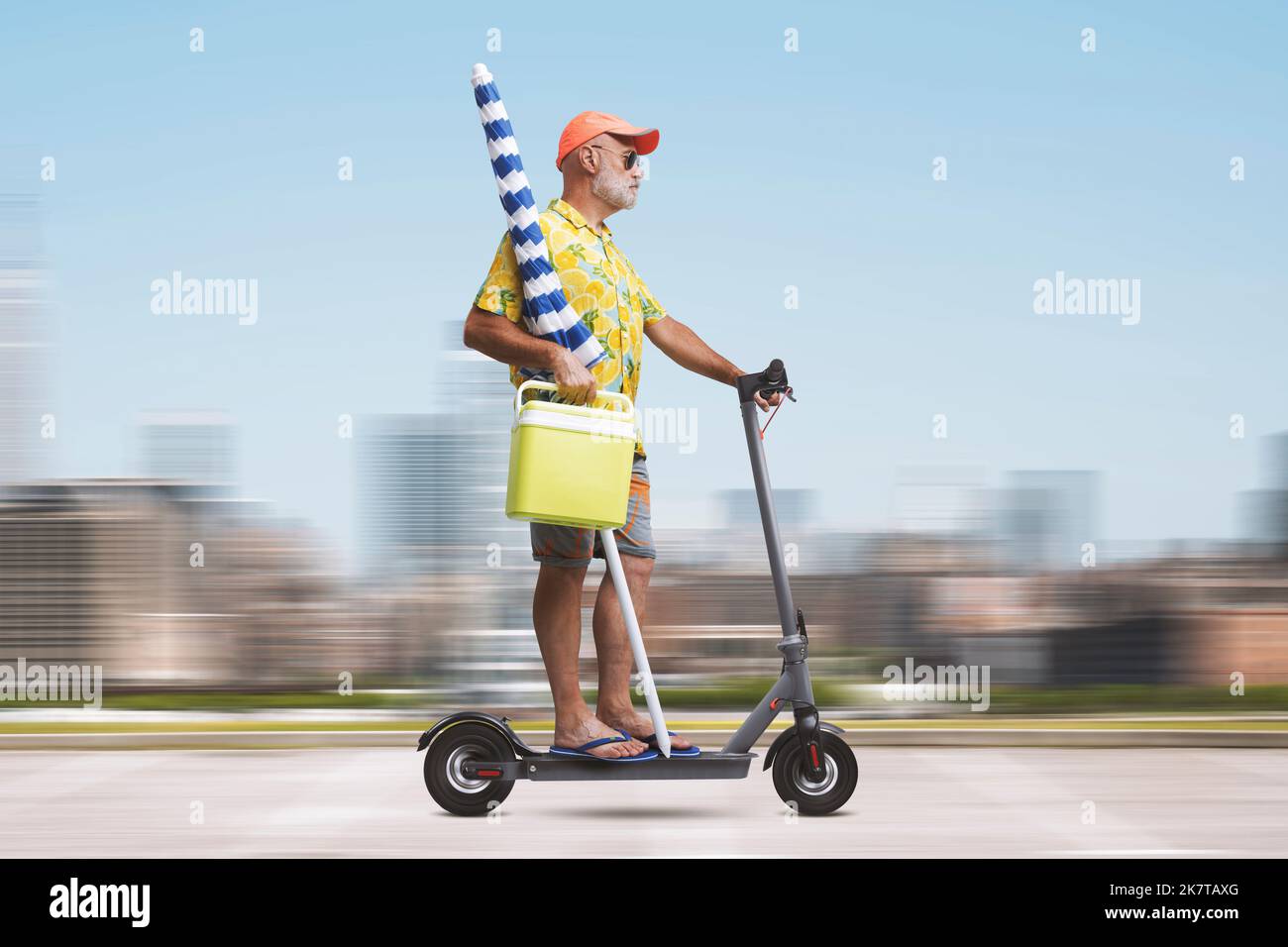Funny senior man holding a beach umbrella and riding an electric scooter, he is going to the beach Stock Photo