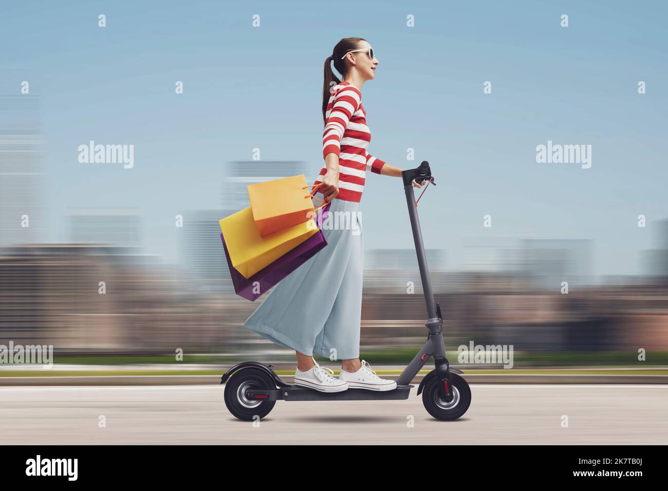 Happy young woman carrying many shopping bags and riding an electric scooter, cityscape in the background, sale and shopping concept Stock Photo