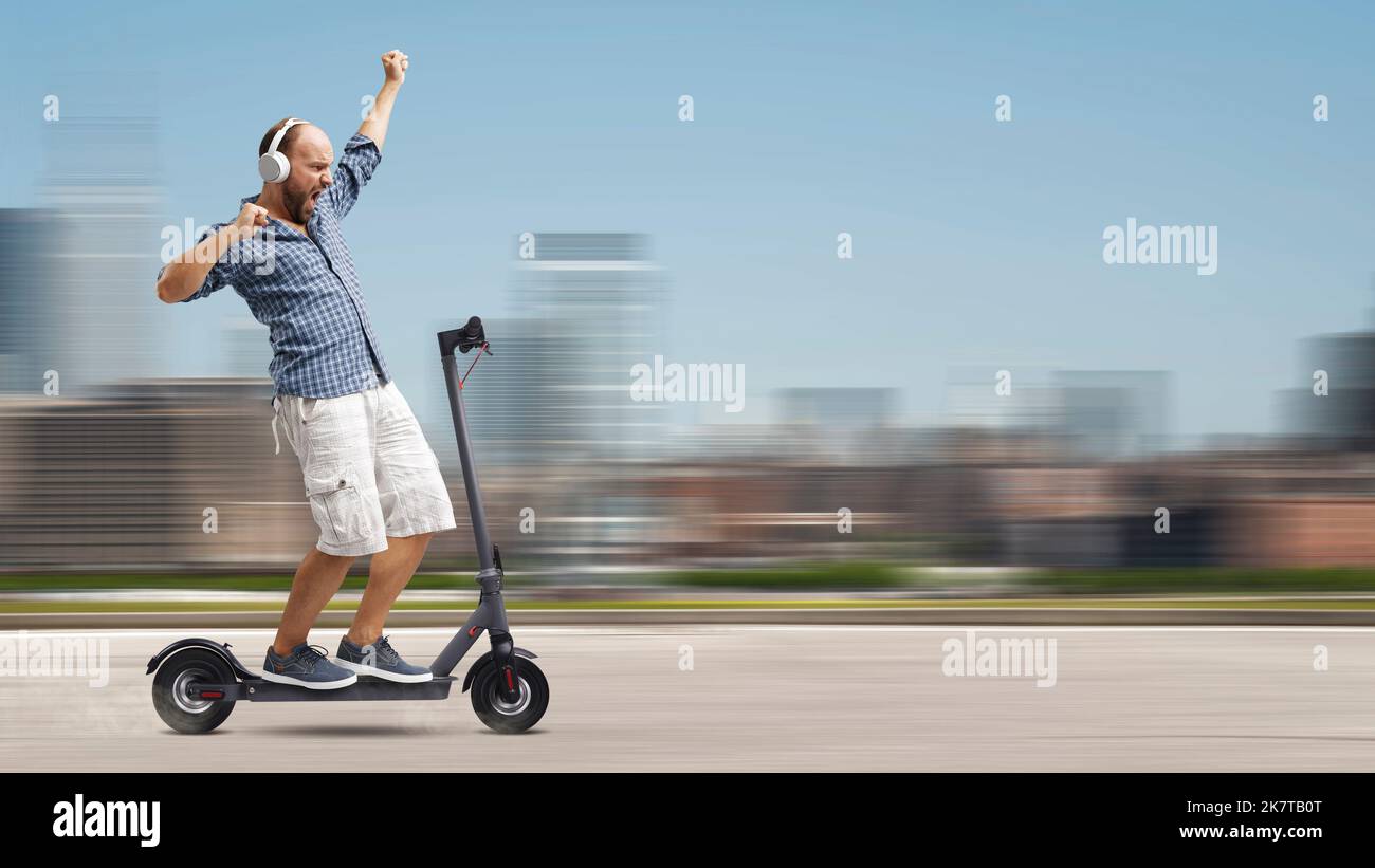 Cheerful man listening to music and dancing while riding an electric scooter, city in the background Stock Photo