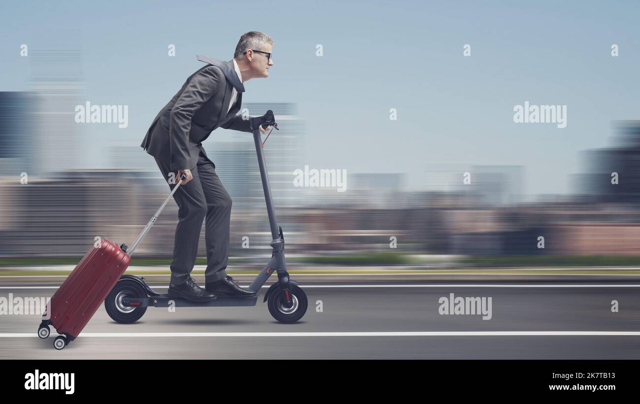 Corporate businessman riding an eco-friendly electric scooter and carrying a roller suitcase, city in the background, sustainable mobility concept Stock Photo