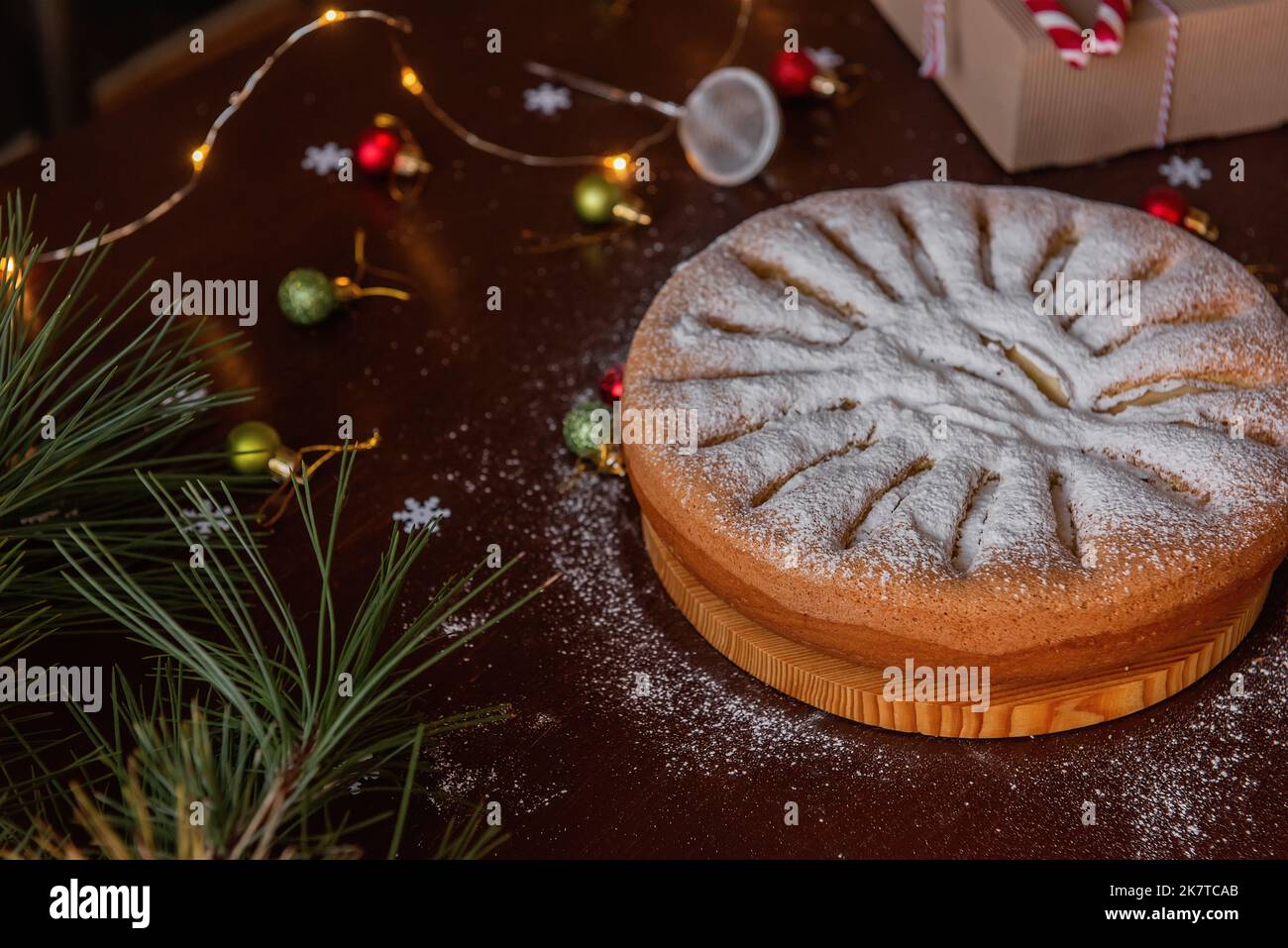 On a wooden brown table lies an apple charlotte sprinkled with powdered sugar. Flat lay top view. Green branch of a pine tree lies next to the pie, a Stock Photo