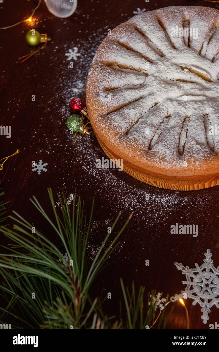 On a wooden brown table lies an apple charlotte sprinkled with powdered sugar. Flat lay top view. Green branch of a pine tree lies next to the pie, a Stock Photo