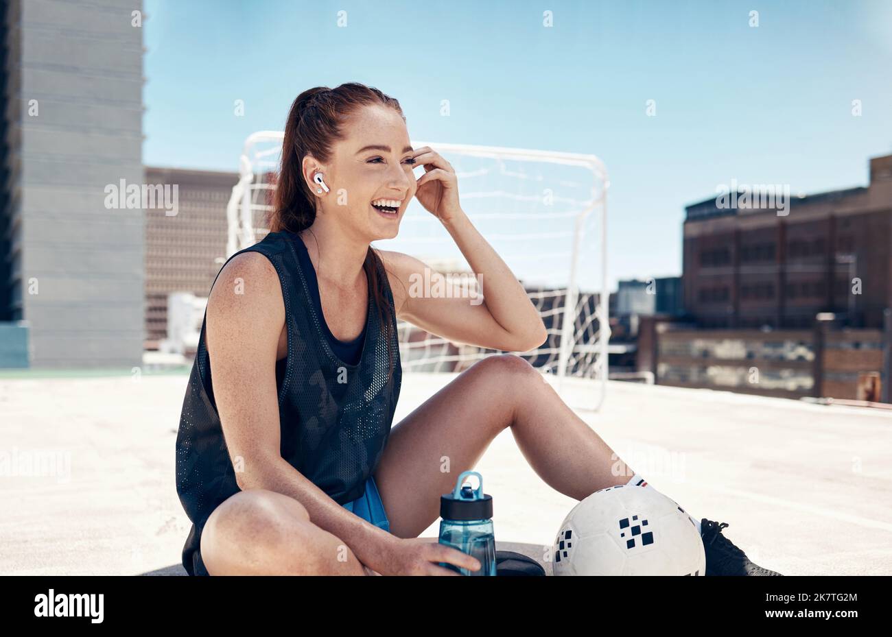 City sports, fitness and woman sitting to relax with water, ball and earbuds. Workout, rest and happy rooftop exercise, fun with urban soccer training Stock Photo