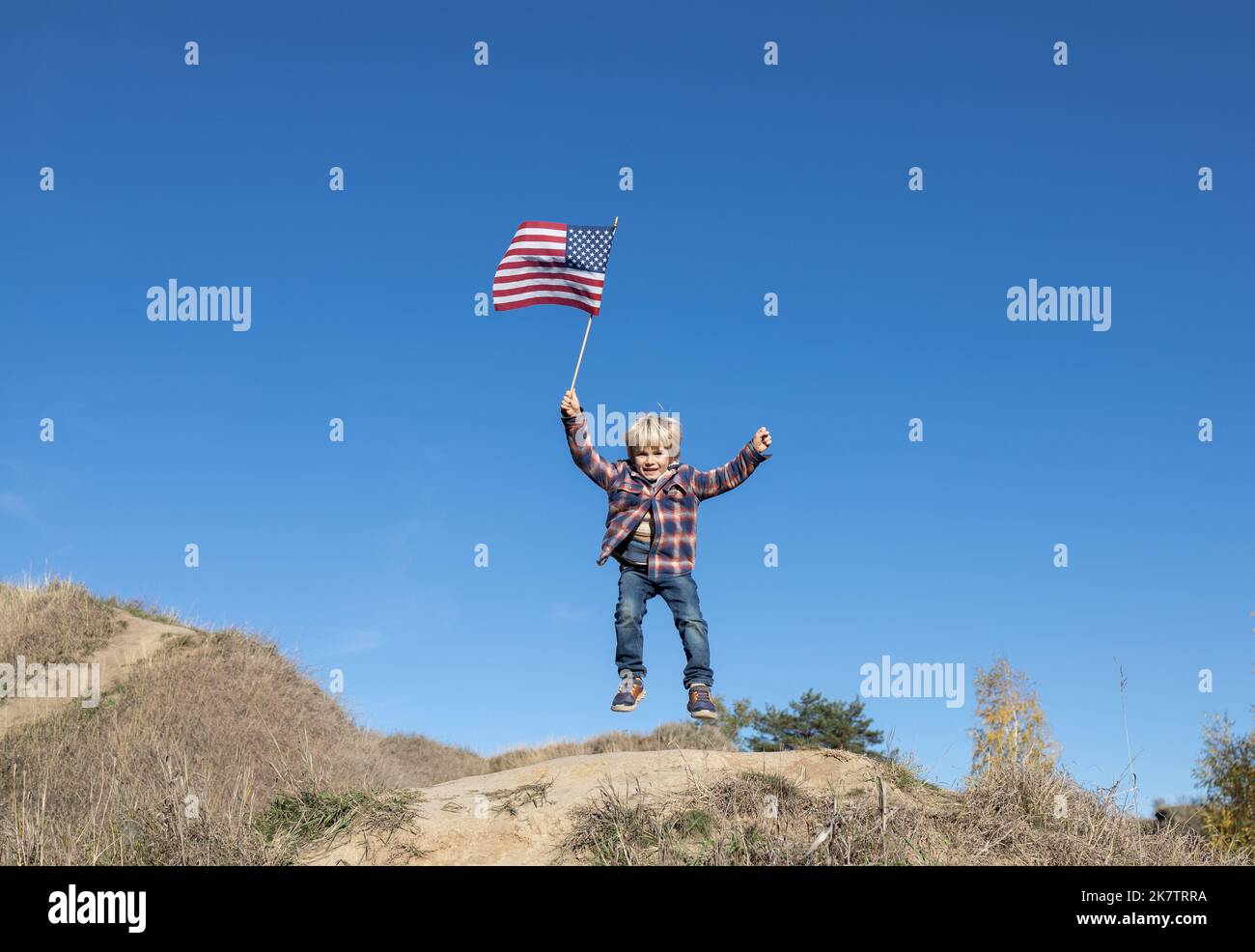 cheerful active little boy joyfully twitching and waving american flag against cloudless blue sky celebrating 4th of july. Independence Day of the Uni Stock Photo