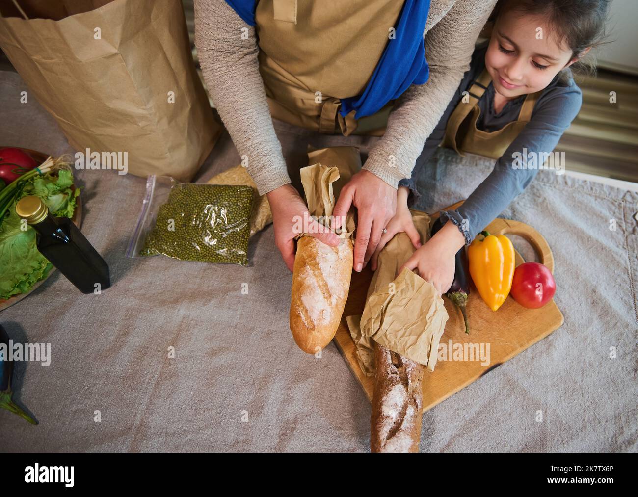 Overhead view of woman with little daughter unpacking food, and loaves of bread from grocery bag together in the kitchen Stock Photo
