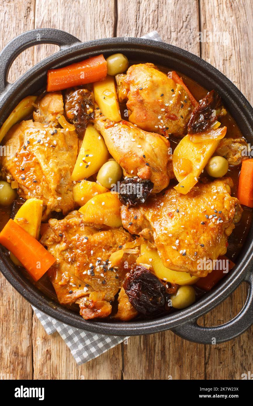 Braised chicken meat with spicy sauce, prunes, carrots, potatoes, olives and onions close-up in a frying pan on a wooden table. Vertical top view from Stock Photo