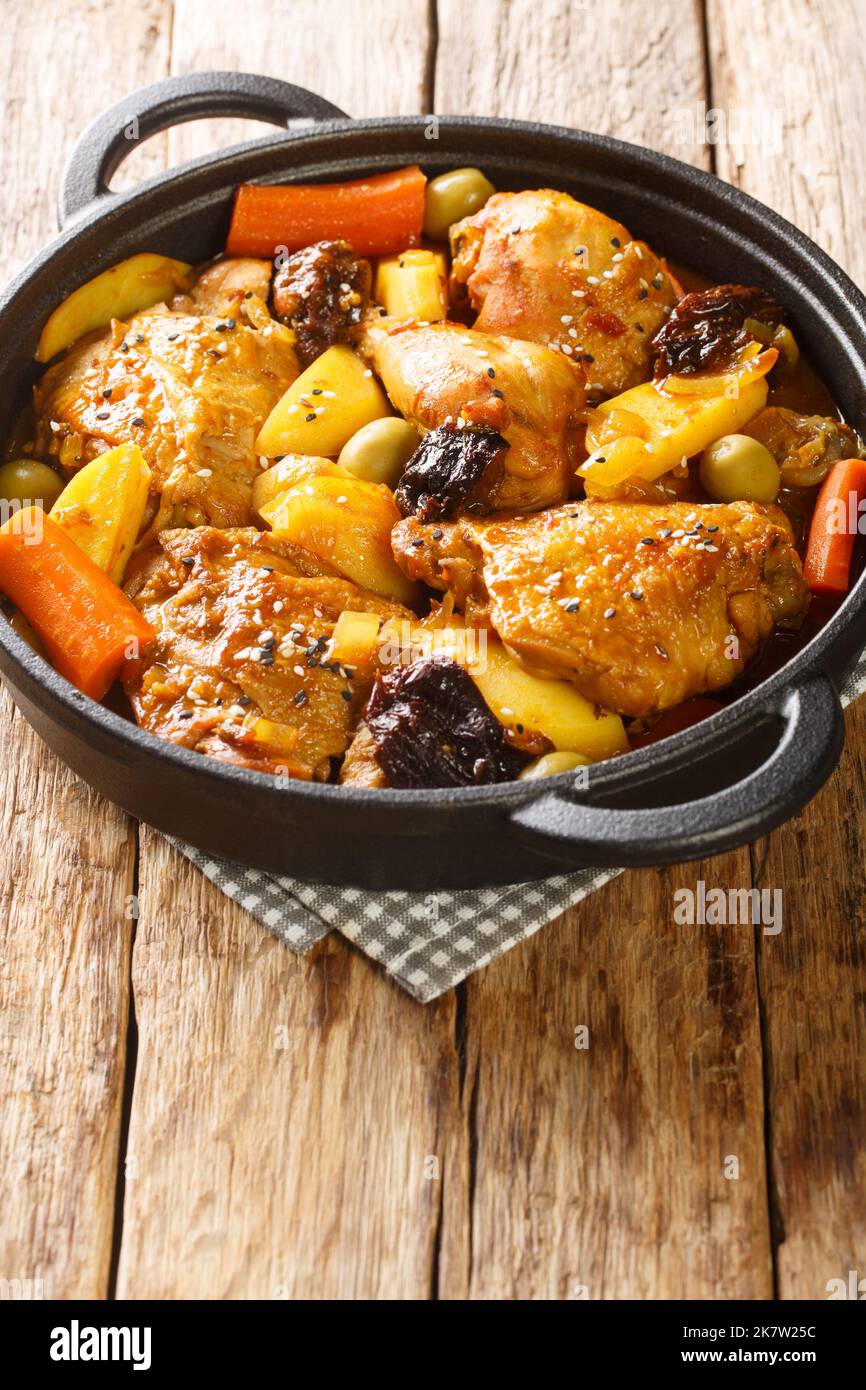 Braised chicken meat with spicy sauce, prunes, carrots, potatoes, olives and onions close-up in a frying pan on a wooden table. Vertical Stock Photo
