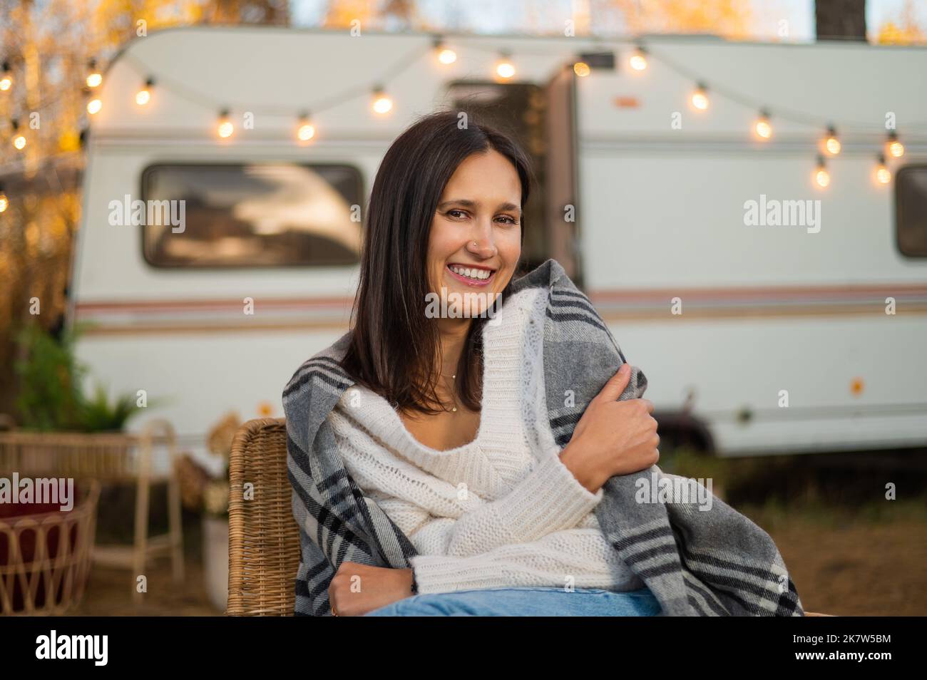 Caucasian woman sits in a wicker chair wrapped in a blanket in the yard near the trailer in autumn.  Stock Photo