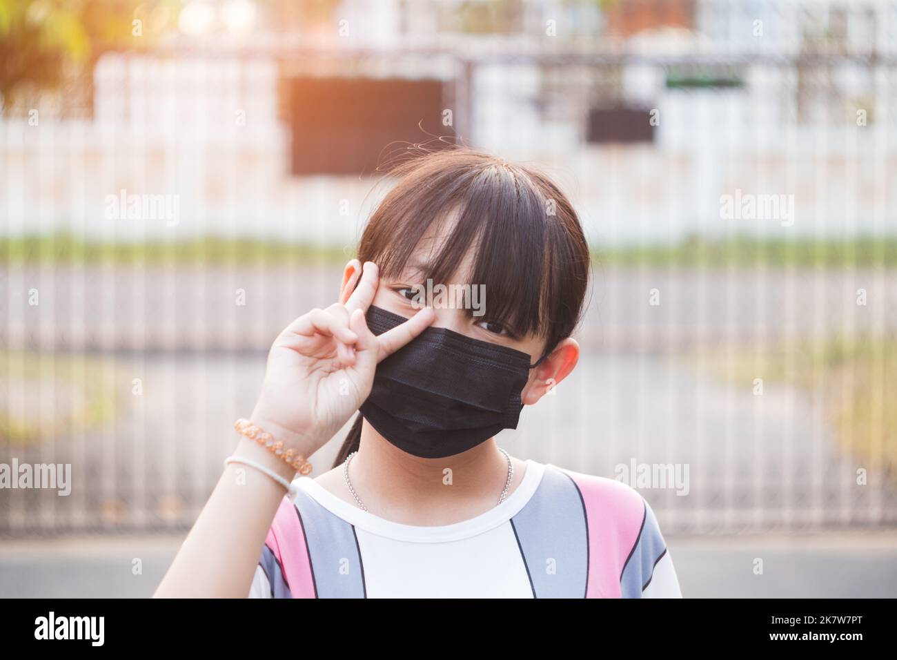 Little girl in a black medical mask smiles and shows thumb up on a pink ...
