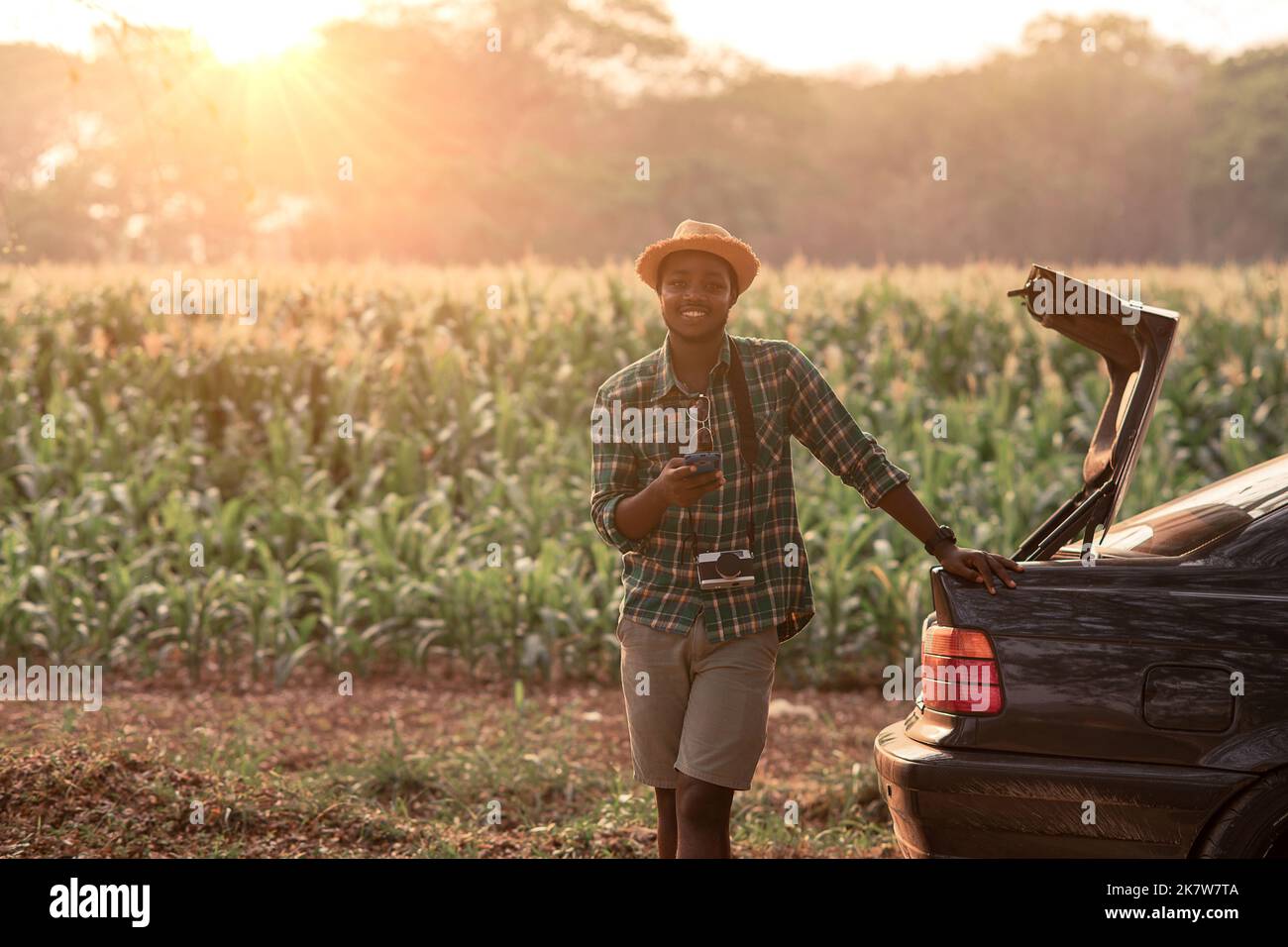 African traveler stands and uses a mobile phone beside his car parked beside the road in an agricultural area.Concept of Adventure travel and transpor Stock Photo