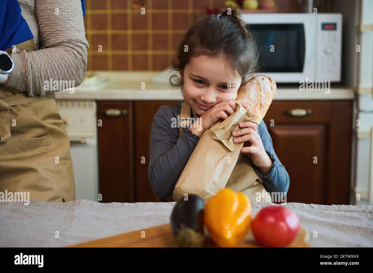 Adorable baby girl holds a hot baguette of freshly baked whole grain sourdough bread, in the kitchen with rustic design Stock Photo