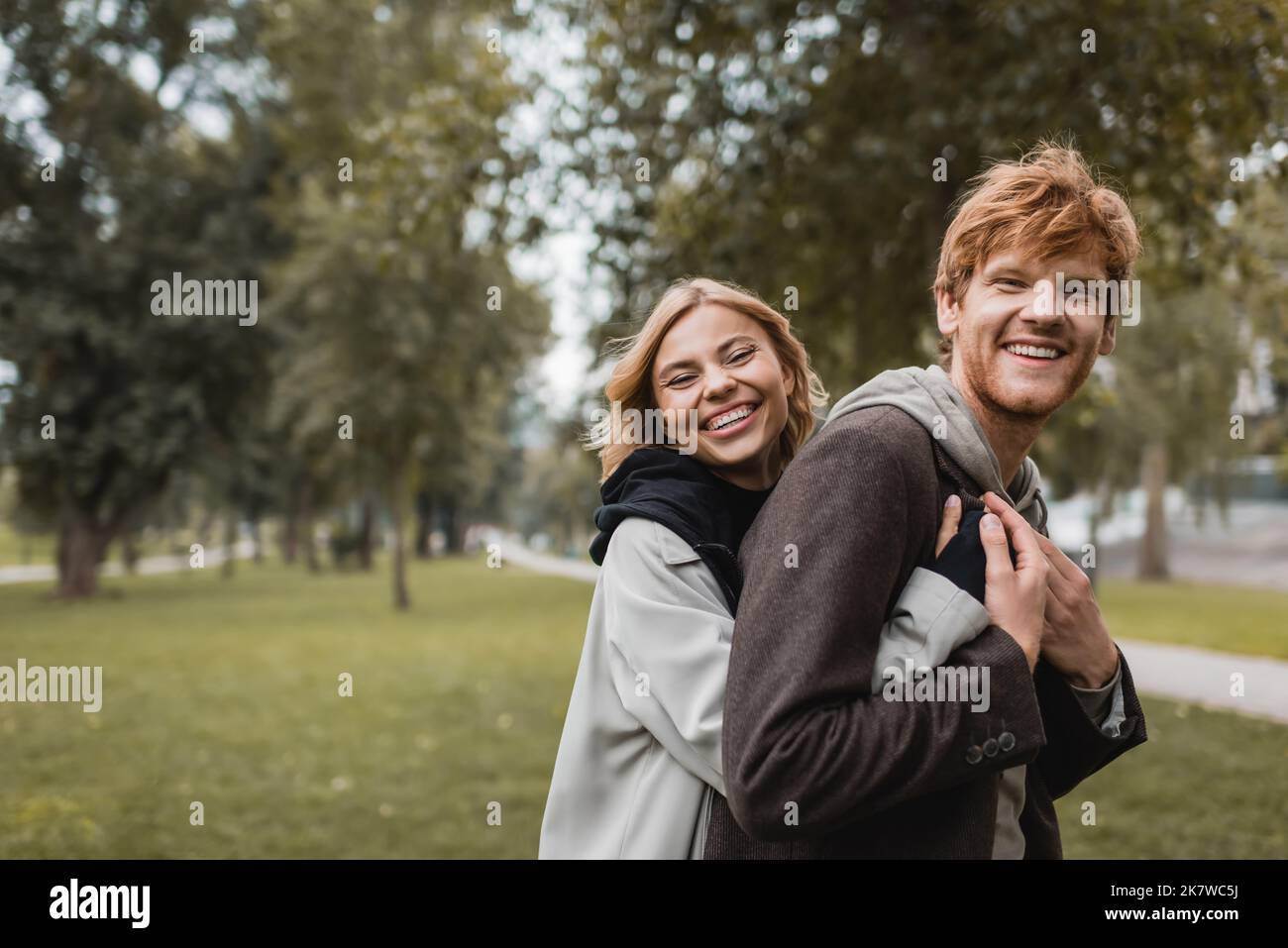 positive young woman in coat hugging cheerful redhead boyfriend in park ...
