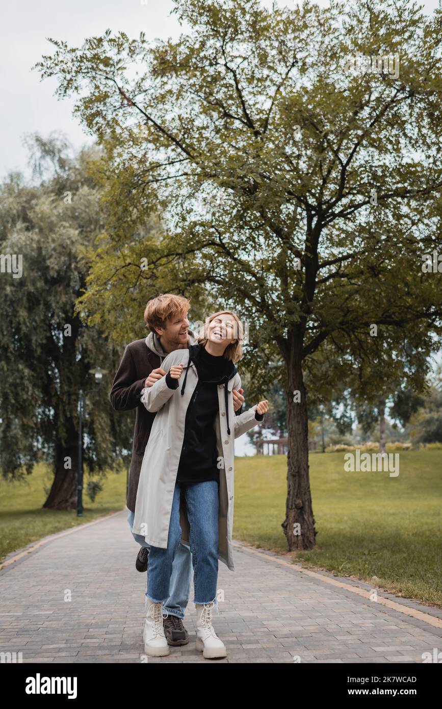 happy man in coat hugging cheerful young woman with closed eyes ...