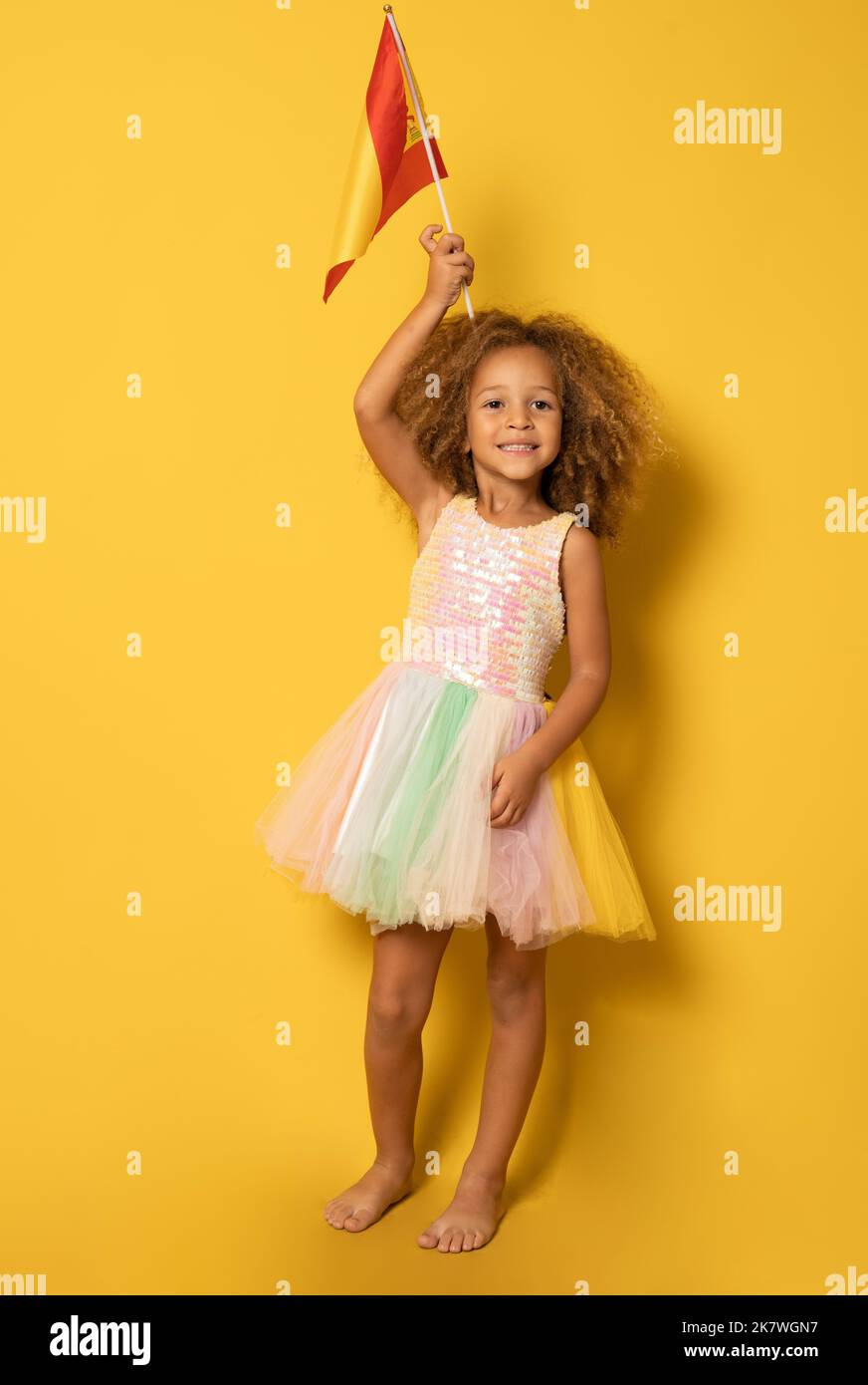 Cute little girl holding Spanish flag standing isolated over yellow background. Stock Photo