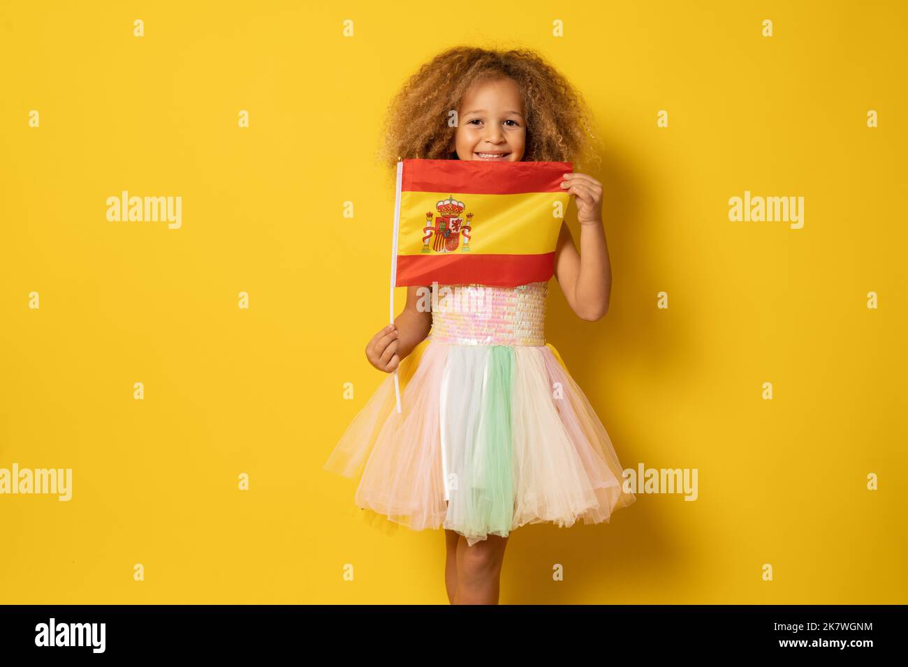 Cute little girl holding Spanish flag standing isolated over yellow background. Stock Photo
