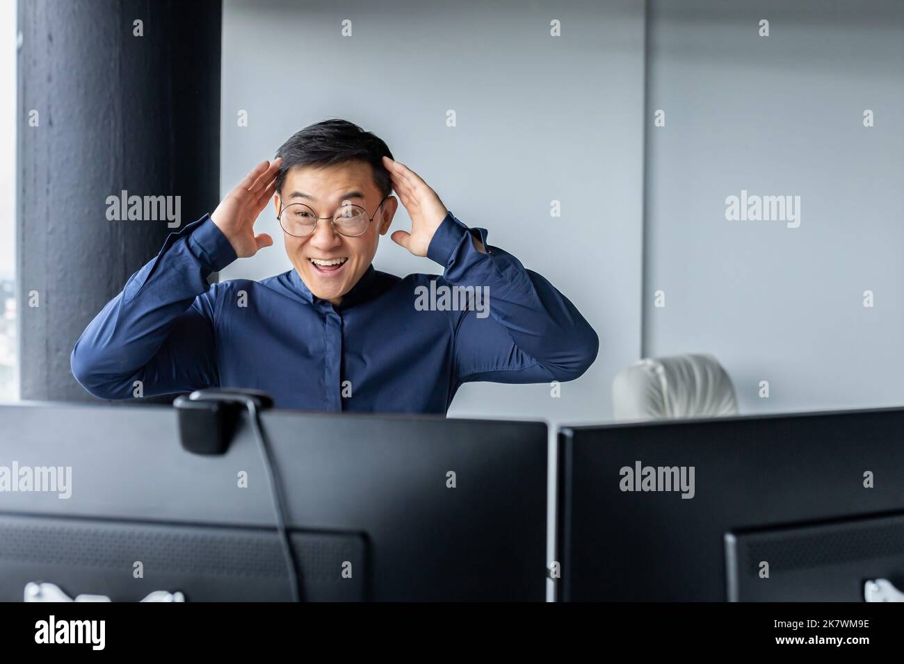 Happy business broker looking at two paired computer monitors and happy, businessman working inside modern office building, asian man celebrating successful deal and signed contract. Stock Photo