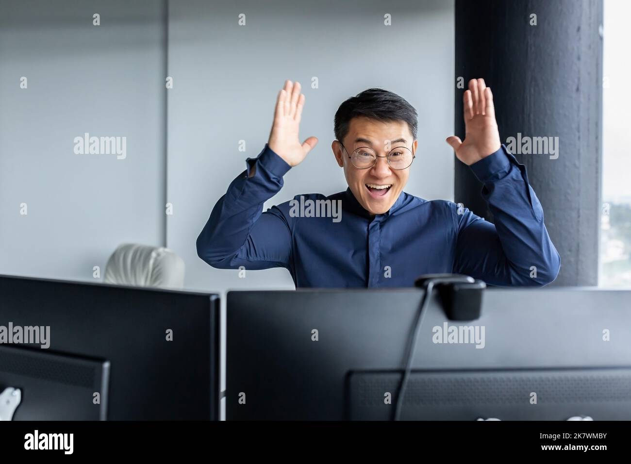 Happy business broker looking at two paired computer monitors and happy, businessman working inside modern office building, asian man celebrating successful deal and signed contract. Stock Photo