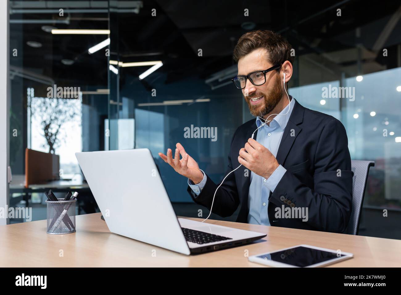 Video call successful businessman boss beard in business suit on online meeting, talking with colleagues using headset and laptop, for remote conversation, man working inside modern office building. Stock Photo