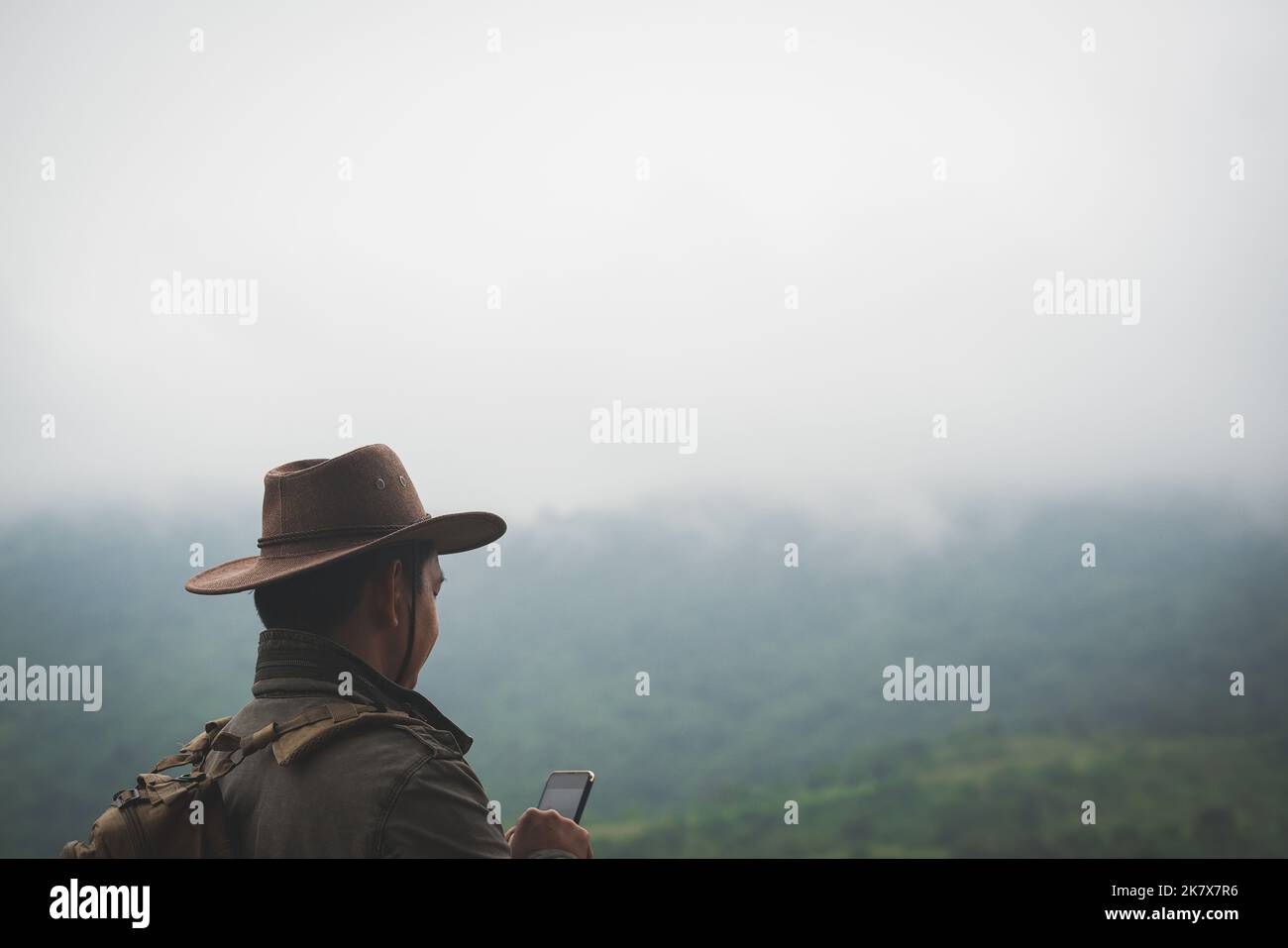 Freedom traveler man in hat carrying a backpack stands at the top of a mountain and using a smartphone on a foggy day.Adventure travel and success con Stock Photo