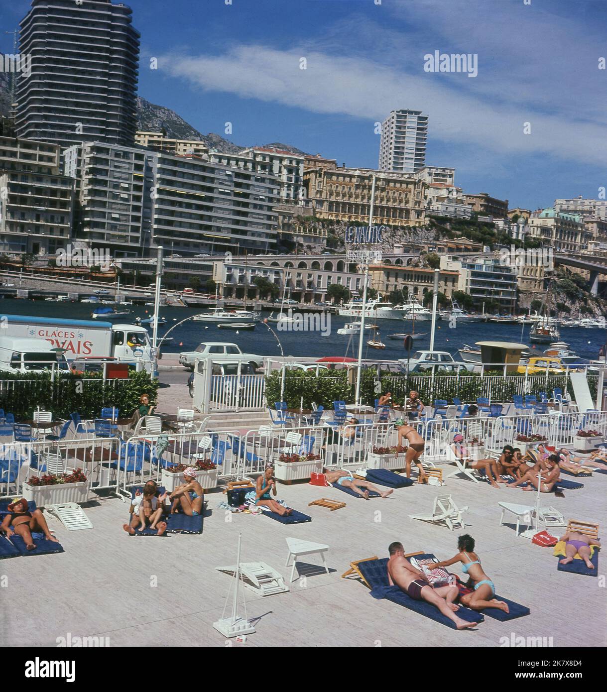 1960s, historical, people sunbathing pool side at the State Nautique Rainier III, an open-air, swimming pool located at Port Hercules, Monaco. The Olympic size, seawater pool opened in 1961. Beside it, restuarant Le Nautic and in the distance, tall buildings, housing luxury apartments overlooking the port. Stock Photo