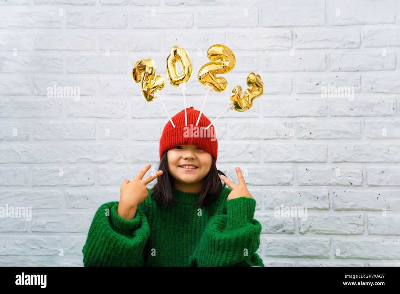 Happy Asian baby girl in a green sweater close-up with golden balloons with numbers 2023 in a hat. Copy space, white brick wall background, girl looki Stock Photo