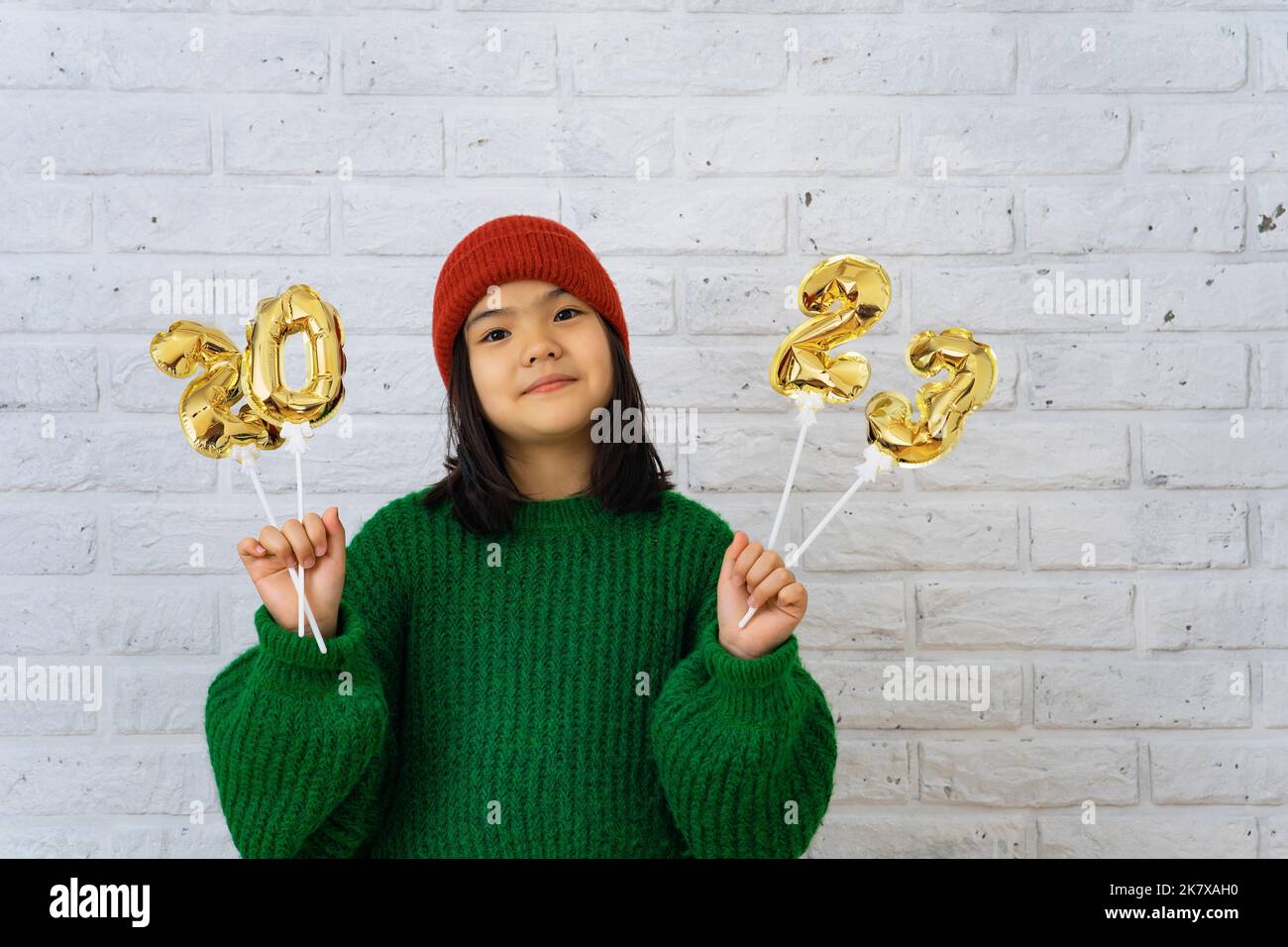 Asian cute girl in red cap and green sweater holding foil balloons with numbers 2023. Copy space, brick white wall background Stock Photo