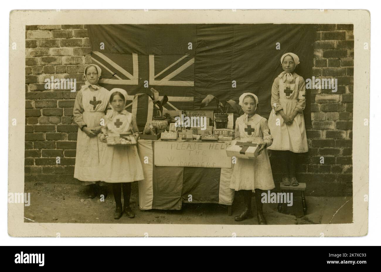 Original and charming WW1 era postcard of working class children dressed in red cross uniforms, with a stall raffling or selling their toys and games, sweets and potted plants to help raise money for wounded soldiers, Union Jack flag in background, 1914-1918 U.K. Stock Photo