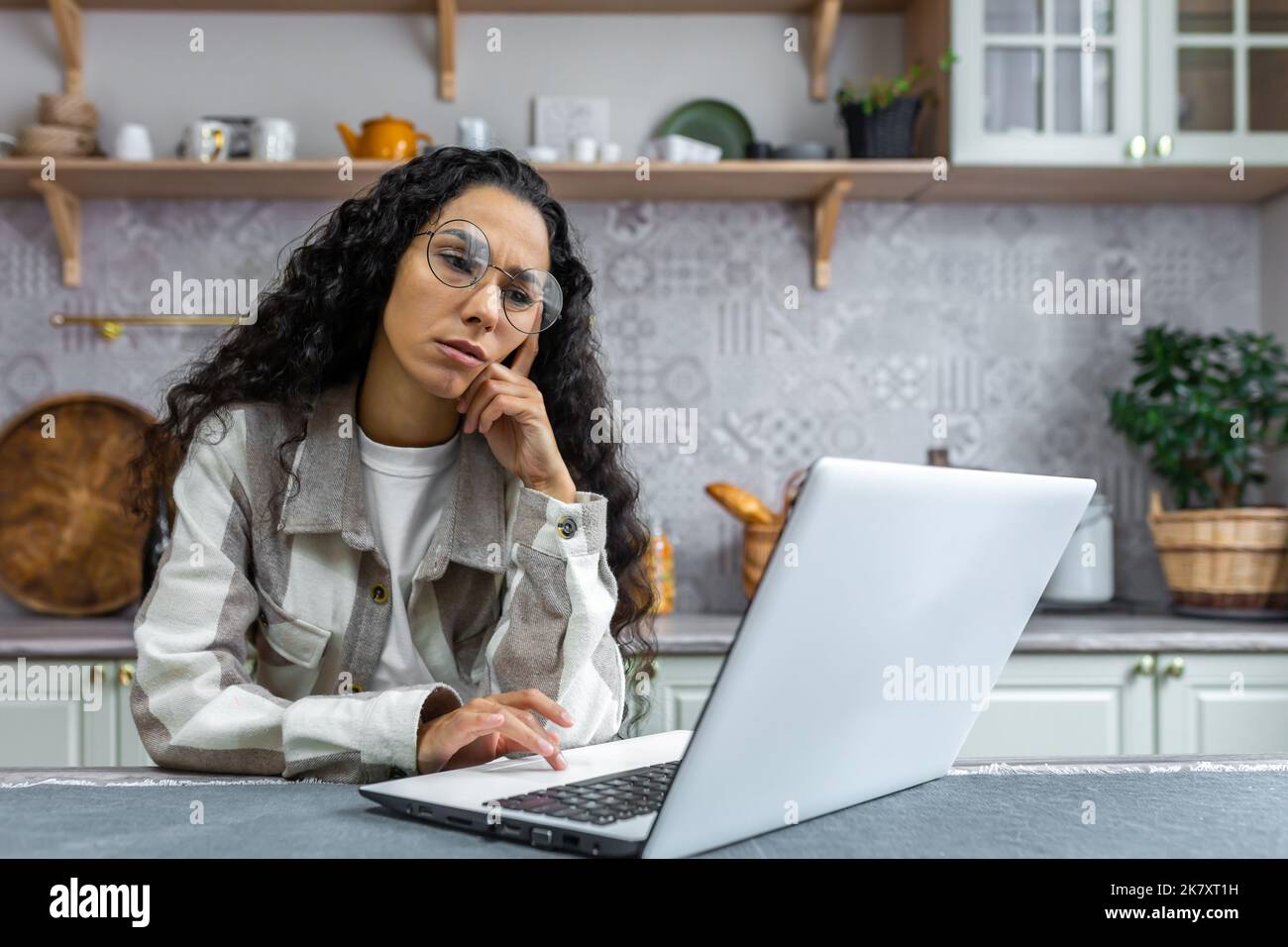 Sad and thinking woman working at home using laptop, Hispanic woman in kitchen at table wearing glasses and curly hair, businesswoman working remotely at home alone. Stock Photo