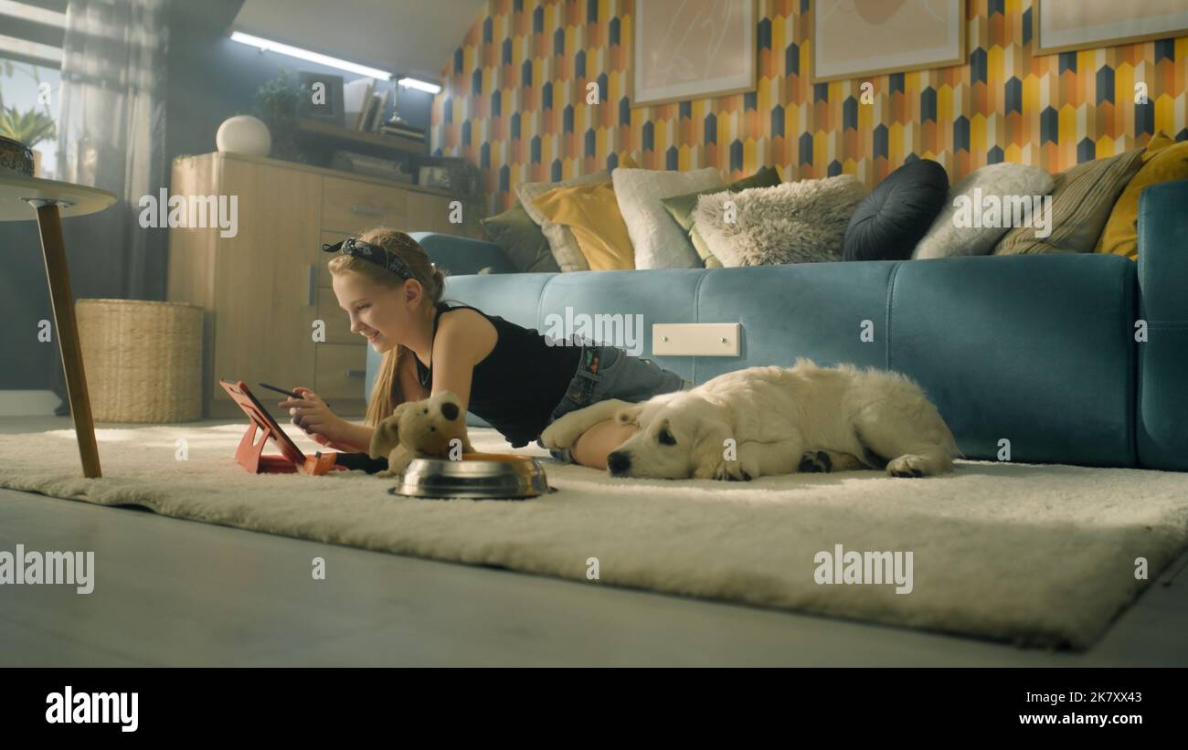 Young girl lying on mild carpet, choosing video on digital tablet computer, dog feeling tired and trying to sleep, spending leisure time at home with pet. Golden retriever. Stock Photo