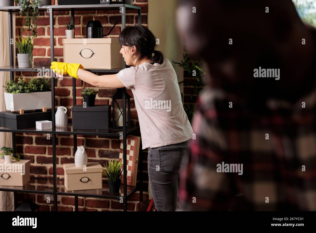 Woman wearing rubber gloves dusting shelves in urban style apartment