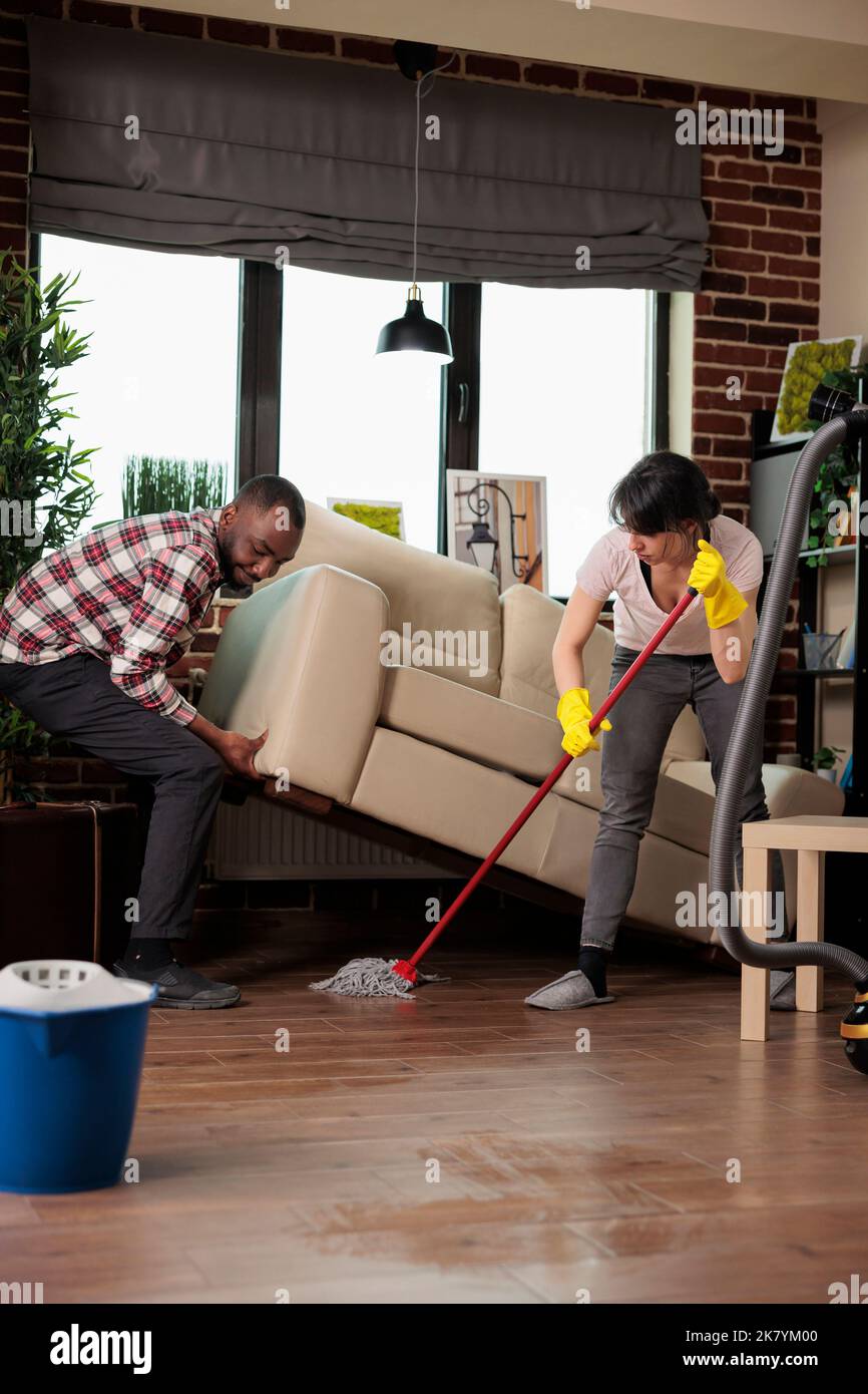 African american man lifts sofa while woman dusts underneath with mop, roommates doing spring cleaning in city apartment. Multiracial friends taking care of keeping home clean and tidy. Stock Photo