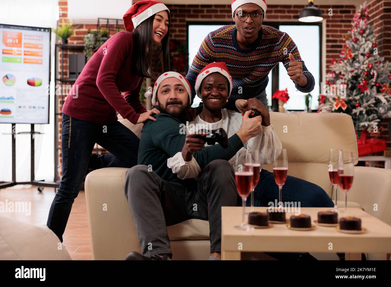 Happy coworkers having fun with video games at event in festive office decorated with xmas tree and ornaments. Playing competition on console and celebrating christmas holiday festivity. Stock Photo