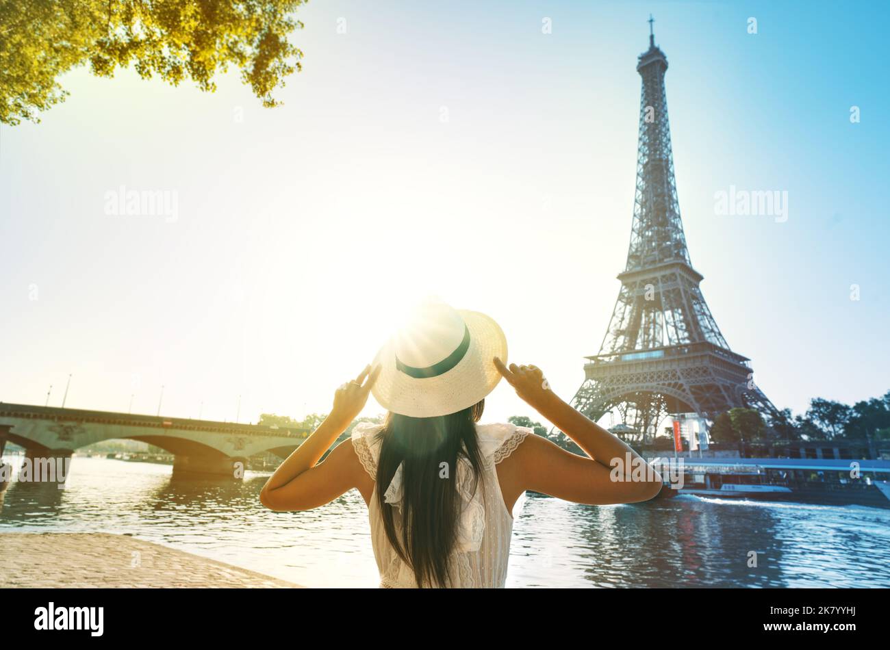 Young woman tourist in sun hat and white dress standing in front of Eiffel Tower in Paris at sunset. Travel in France, tourism concept Stock Photo