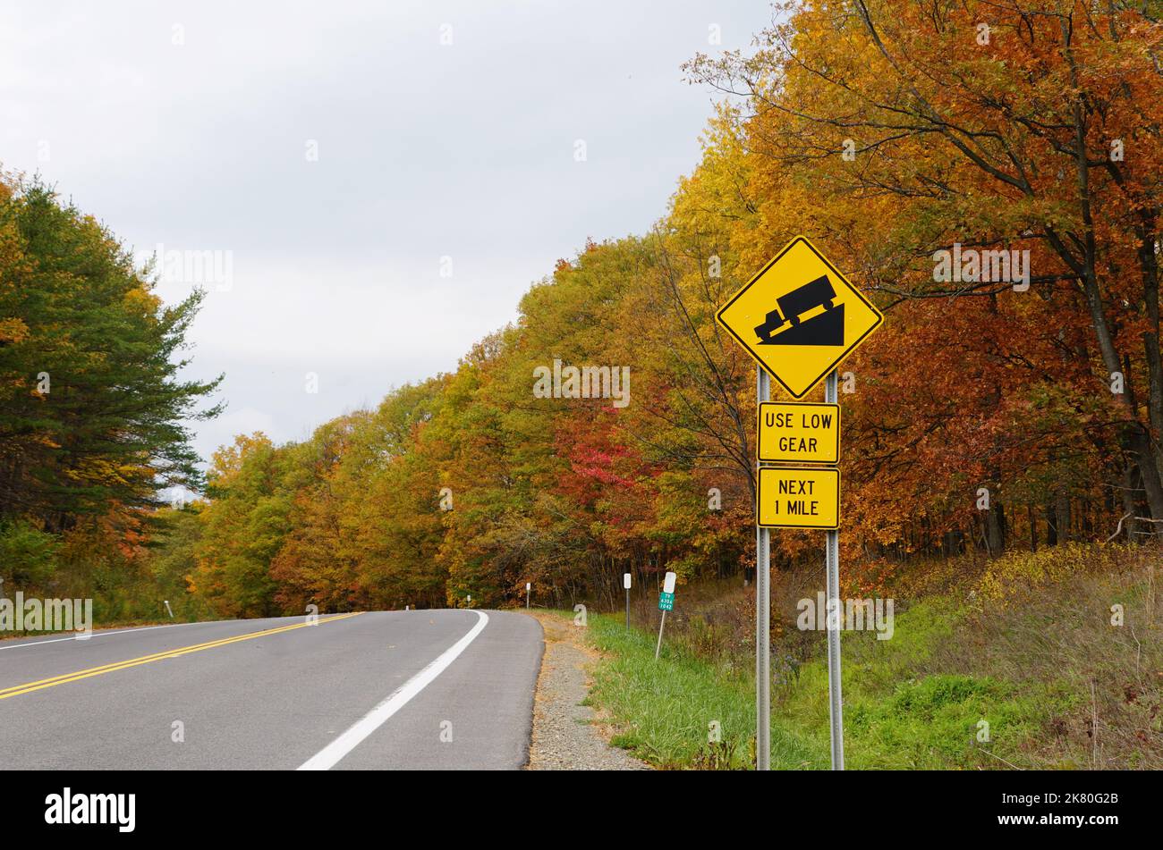 Upstate New York, U.S.A - October 17, 2022 - A road with sharp corner and sign for trucks to use low gear, with the background of fall foliage Stock Photo