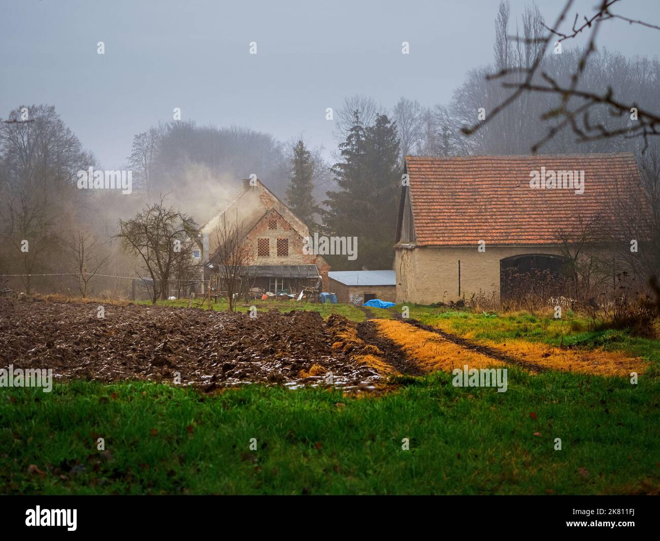 plowed field covered with a snow and old farmhouse on a background in an old village in a rainy misty winter day Stock Photo