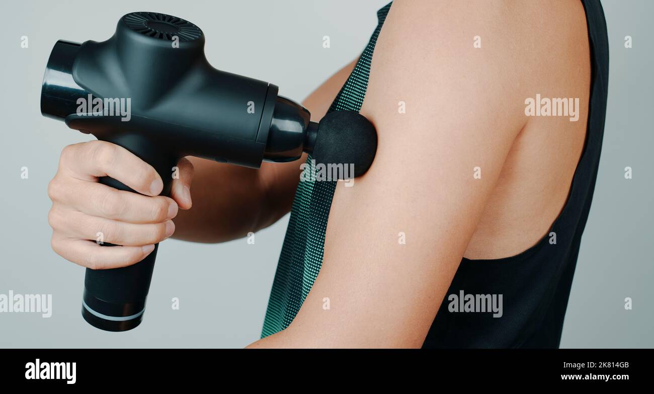 a young caucasian man, wearing sports clothes, massages the muscles of his arm with a massage gun Stock Photo