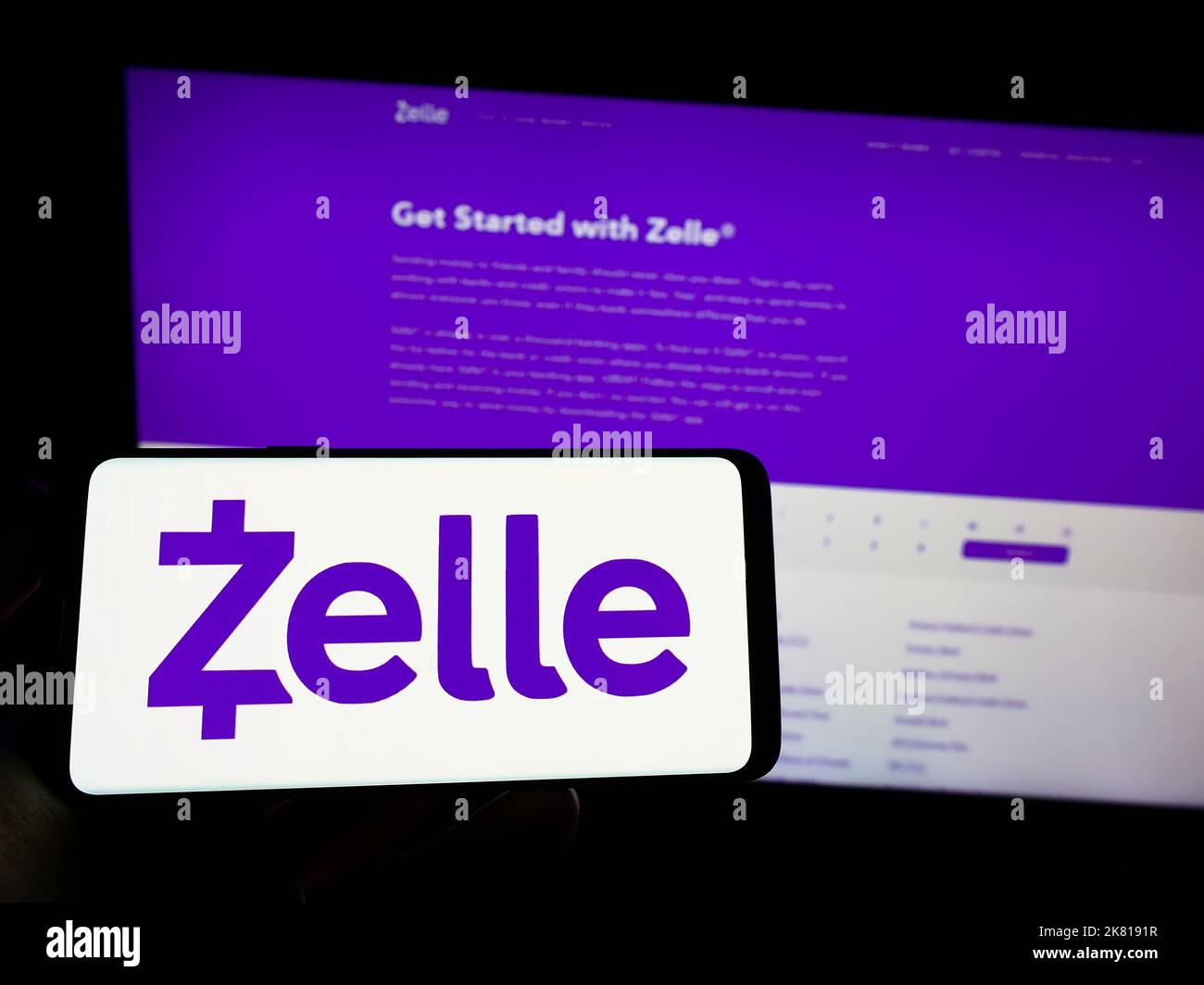 Person holding mobile phone with logo of American digital payments company Zelle on screen in front of business web page. Focus on phone display. Stock Photo