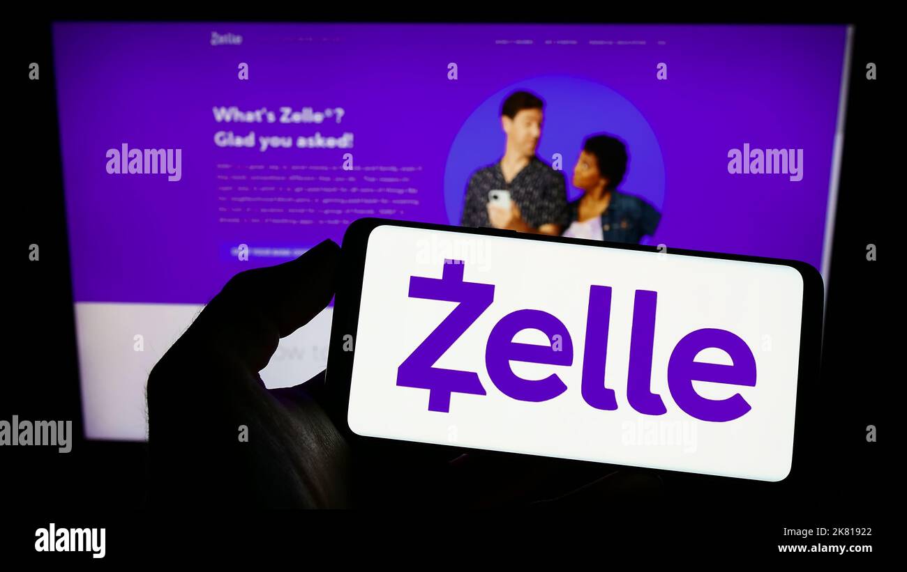 Person holding cellphone with logo of US digital payments company Zelle on screen in front of business webpage. Focus on phone display. Stock Photo