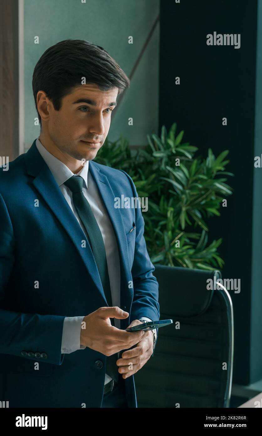 Businessman in suit with mobile phone in hands. Man typing message, using social net. Checking news. Making business meeting. Modern office.General ma Stock Photo