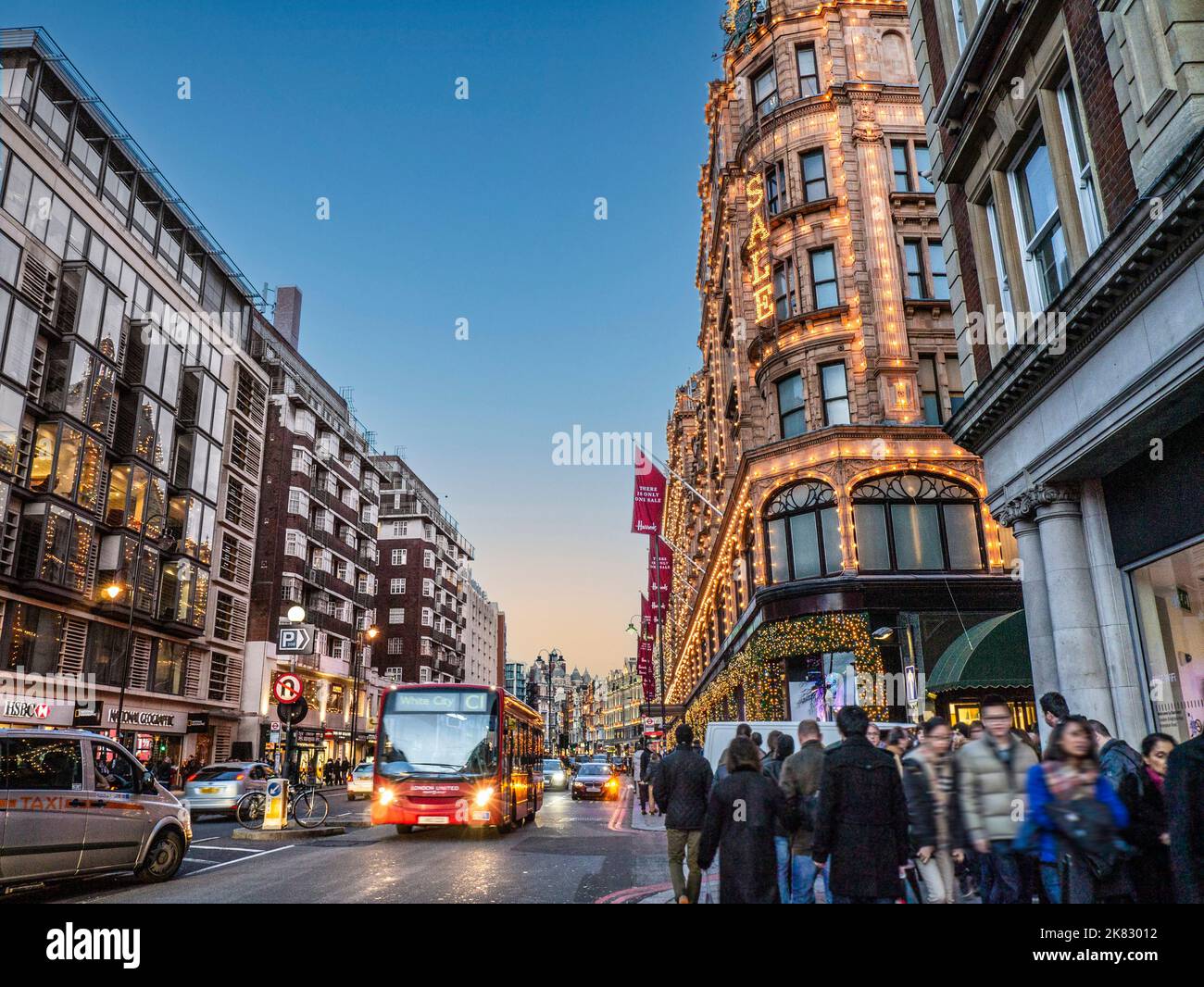 BROMPTON ROAD KNIGHTSBRIDGE WINTER SALES with red bus and Harrods department store behind at dusk night with lit 'Sale' sign busy shoppers crowds traffic sunset Harrods Knightsbridge London SW1 Stock Photo