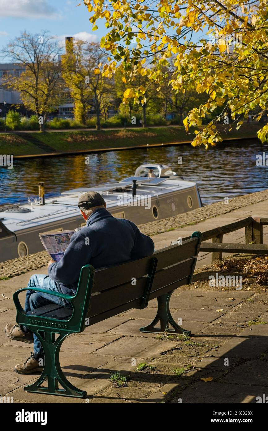 Candid view of a mature man sitting relaxing while reading a newspaper next to the River Ouse along The Dame Judi Dench Walk, York, North Yorkshire,UK. Stock Photo