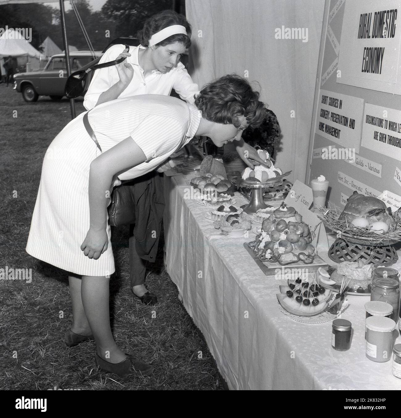 1960s, historical, two young women attending an agricultural show, look at the bread and cakes on an exhibition stand promoting the benefits of the Rural Domestic Economy, England, UK. On the stall, information on the shortcourses available from groups such as the Women's Briitsh legion, and the Womens Guild, on cookery, food preservation and home management. A card says Use of Cream another says 'Chicken baked in a hot water crust'. Stock Photo