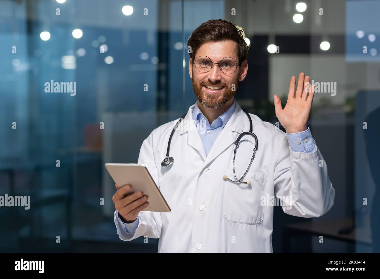 Senior mature doctor with tablet looking at camera and holding hand up greeting gesture, man in white medical coat working inside modern clinic, using tablet computer at work. Stock Photo