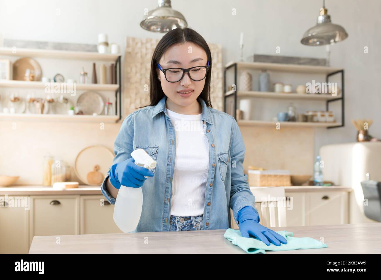 Maid services. Young beautiful Asian woman works as a cleaner. Cleans the house. Wipes the table with a cloth in rubber gloves, sprays the surface with a spray. Stock Photo