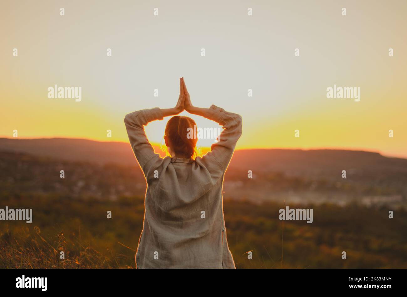 Back view of woman with arms in namaste overhead on hill at sunset Stock Photo