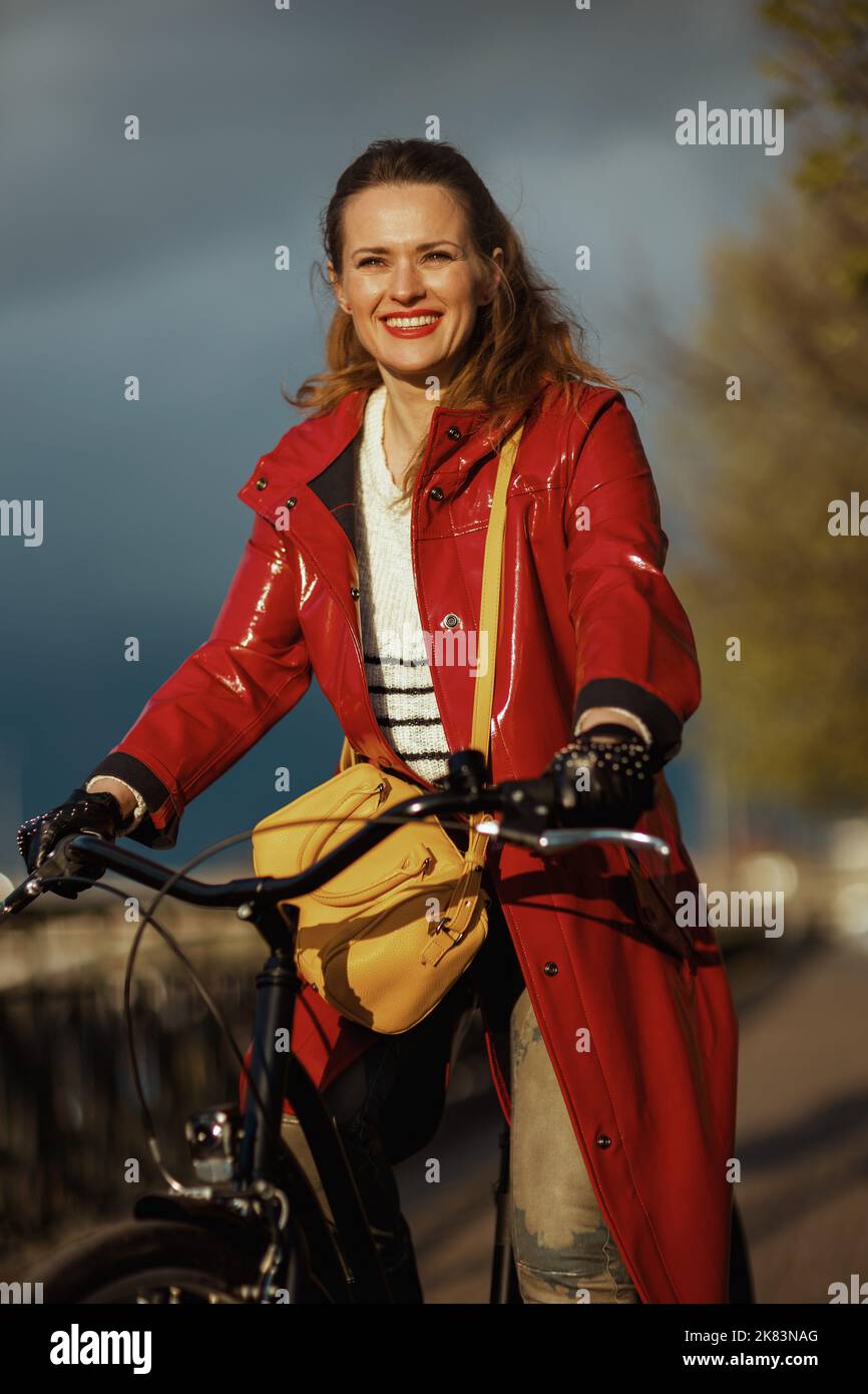 smiling modern 40 years old woman in red rain coat outside on the city ...