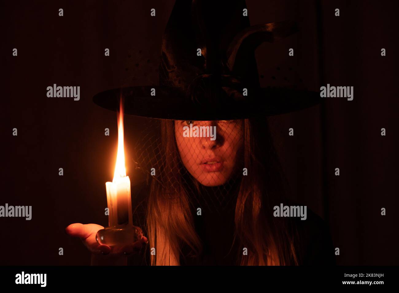 A girl in a witch's hat with a candle in her hands in the dark, a witch and a flame of a switch in the dark, halloween, magic and sorcery Stock Photo