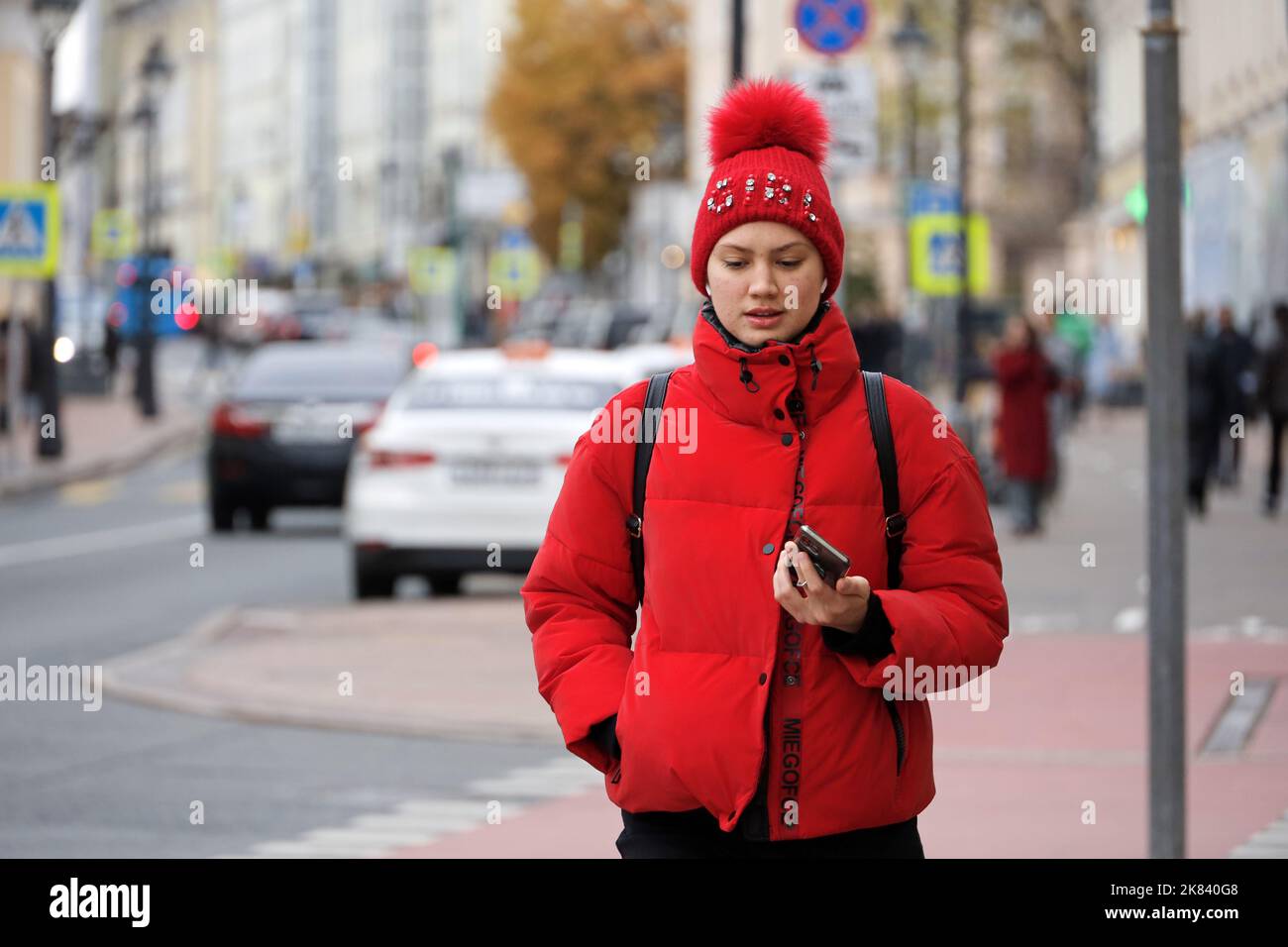 Girl in wireless headphones wearing red jacket and hat talking on smartphone while walking on a street. Using mobile phone in autumn city Stock Photo