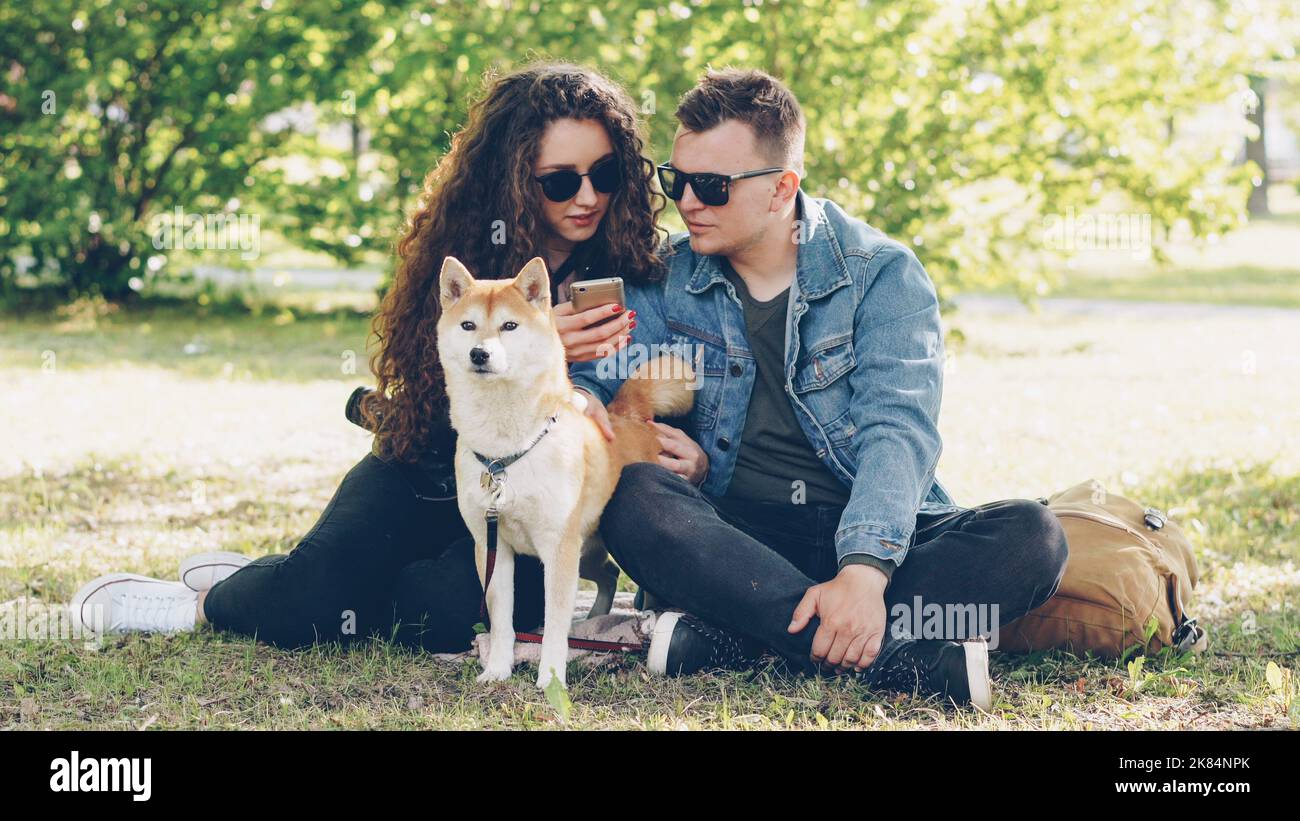 Joyful husband and wife are using smartphone resting on lawn in park, watching screen and talking and fussing dog. Modern technology, domestic animals and city concept. Stock Photo
